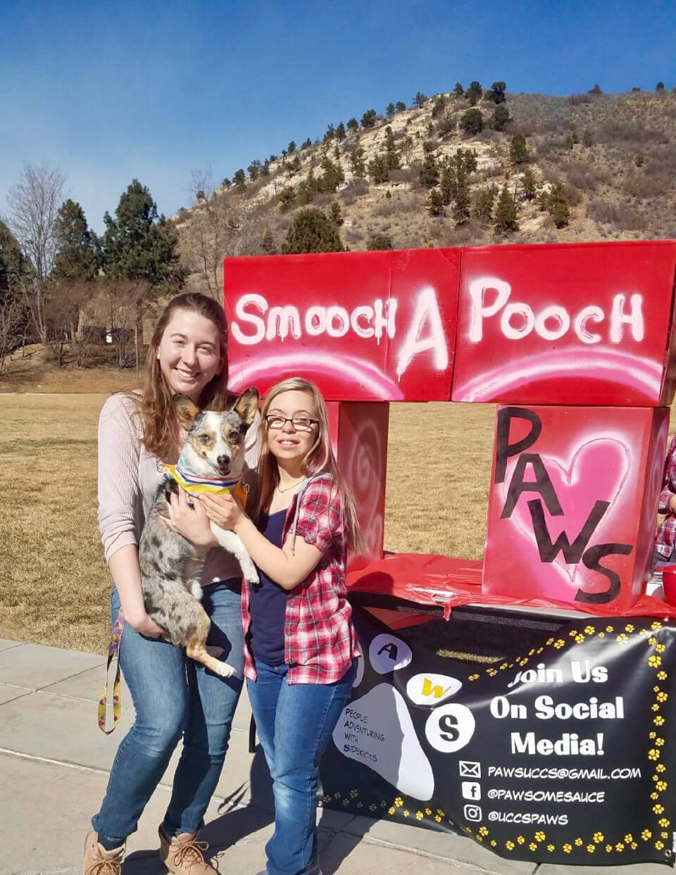 Two students holding a dog in front of a smooch a pooch sign